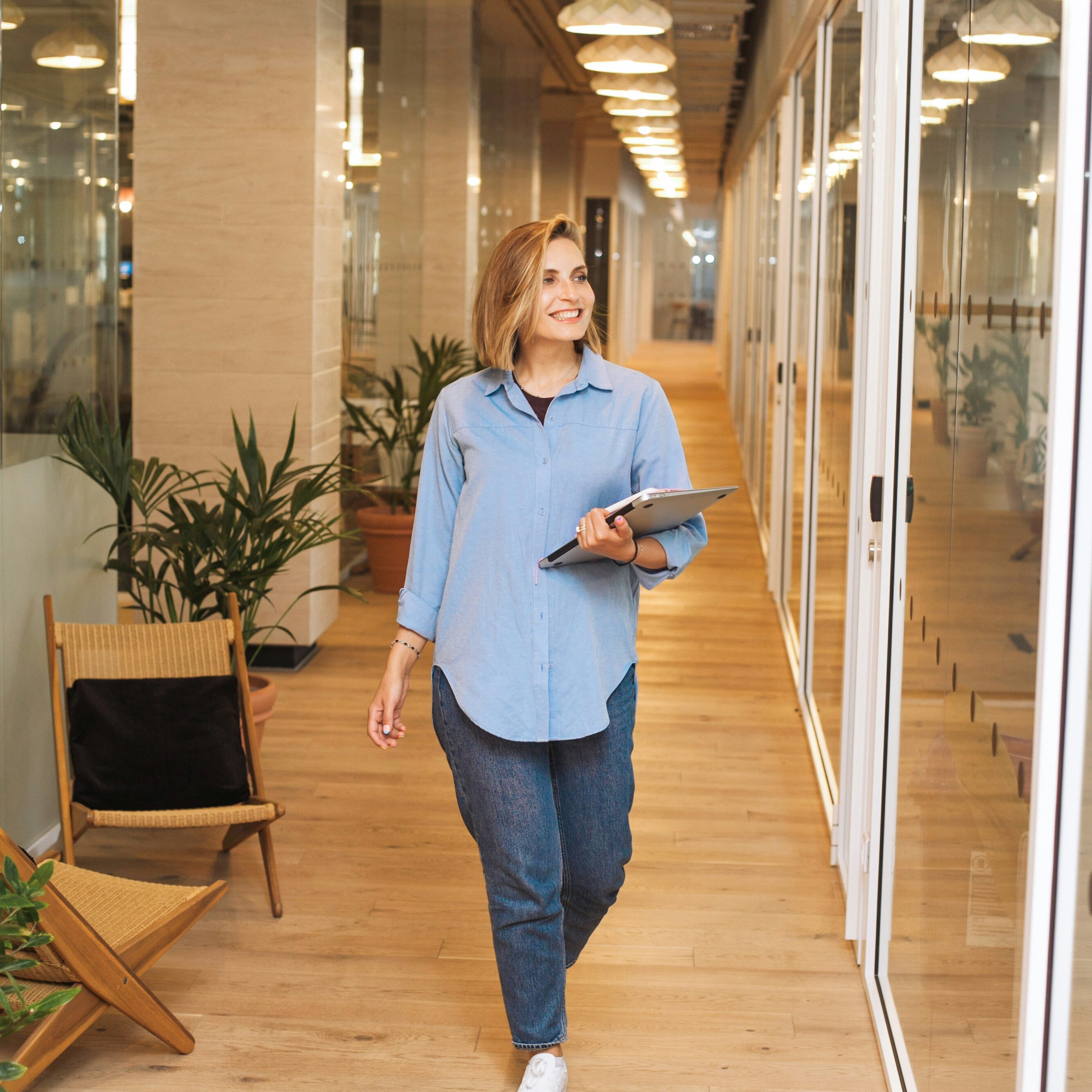 Woman in business attire walking confidently through a brightly lit modern office with glass walls, holding a tablet and a small bag.